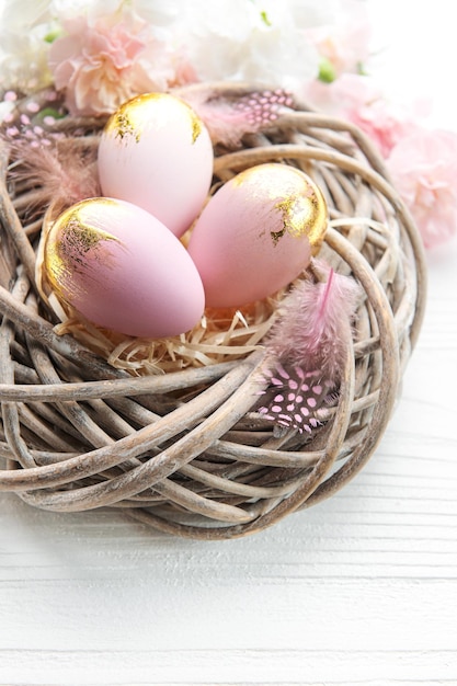 Easter eggs painted pink and gold and feathers in a nest on a white wooden background