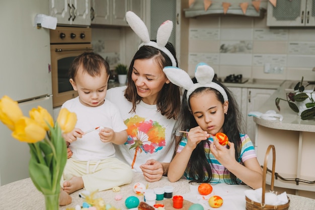 부활절 가족 전통 사랑하는 젊은 어머니는 식탁에 함께 앉아 있는 동안 부활절 달걀을 칠하는 방법을 아이들에게 가르칩니다.
