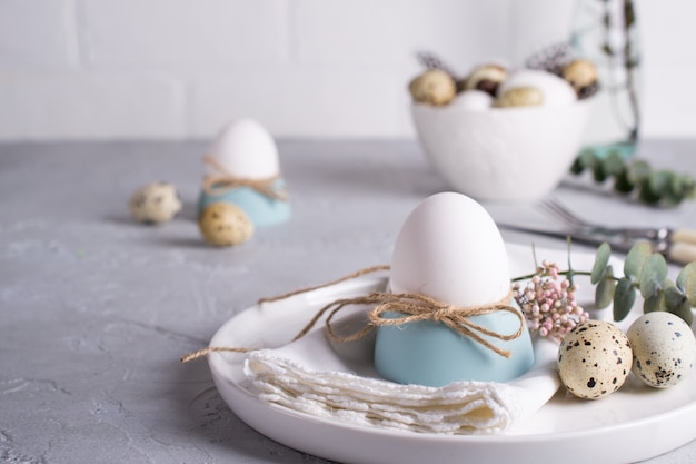 Пасхальная праздничная сервировка с белыми куриными яйцами в чашечках яиц, веточках листьев эвкалипта. ,