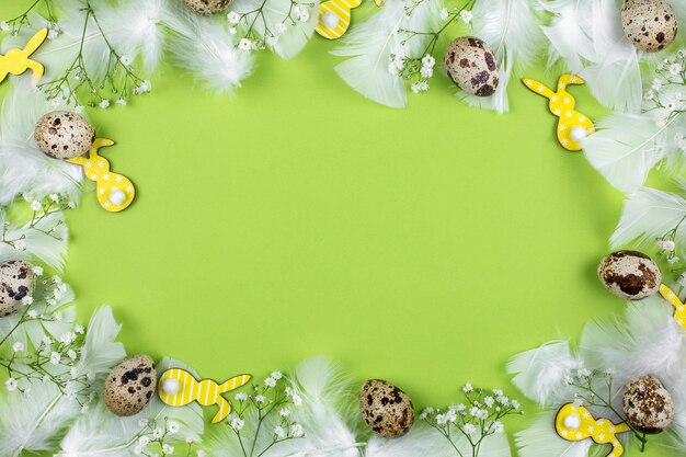 Easter flat lay with quail eggs, easter bunnys, flowers and white feathers on a green background.