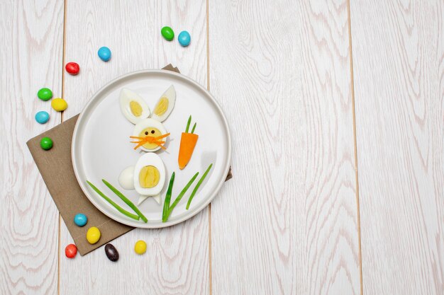 아이들을 위한 부활절 재미 있고 창의적인 건강한 아침 식사 점심 음식 아이디어 접시 흰색 나무 테이블 배경에 삶은 달걀 껍질을 벗긴 당근 채소로 만든 토끼 토끼 평면도 평면 복사 공간
