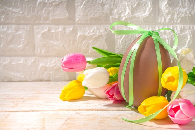 Пасхальная открытка фон с огромным большим шоколадным яйцом, с праздничным букетом цветов тюльпанов ленты, на освещенном солнцем деревянном классическом фоне копией пространства