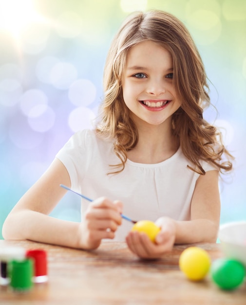 easter, holiday and child concept - happy girl with brush coloring easter eggs over lights background