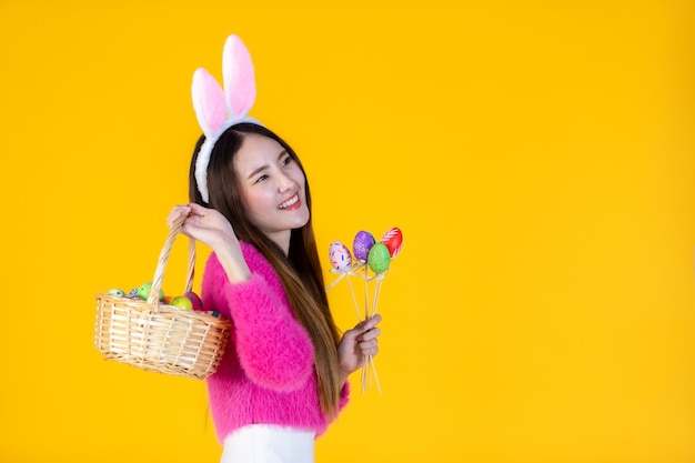 Easter holiday concept, happy asian young woman wearing bunny ears hand holding a basket with colorful easter eggs in isolated on yellow blank copy space studio background.