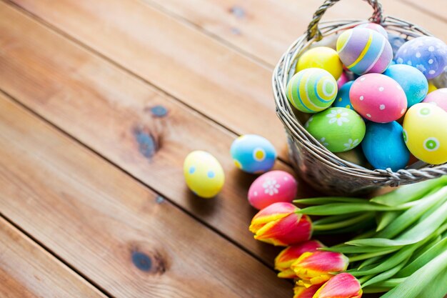 easter, holidays, tradition and object concept - close up of colored easter eggs in basket and tulip flowers on wooden table with copy space