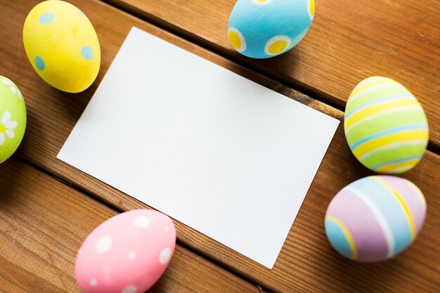 easter, holidays, tradition and object concept - close up of colored easter eggs and blank white paper card on wooden surface with copy space