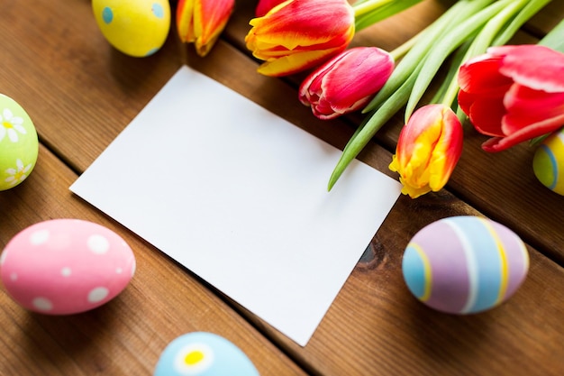 easter, holidays, tradition and object concept - close up of colored easter eggs, tulip flowers and blank white paper card on wooden surface with copy space