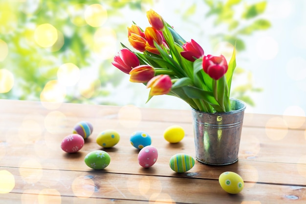 부활절, 공휴일, 전통 및 개체 개념 - 녹색 자연 배경 위에 있는 나무 테이블에 있는 양철 양동이에 색깔 있는 부활절 달걀과 튤립 꽃을 닫습니다.