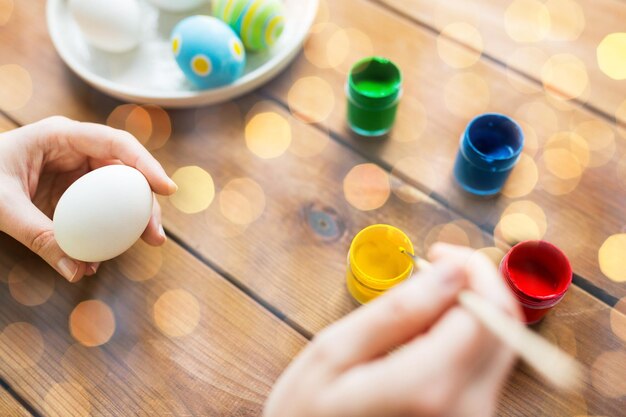 Foto concetto di pasqua, vacanze, tradizione e persone - primo piano delle mani della donna che colorano le uova di pasqua con i colori e spennellano le luci
