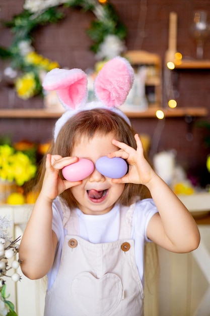 Easter a little child girl plays with Easter eggs covering her face with them in the ears of a hare and smiles at home at the festive table at home in the kitchen