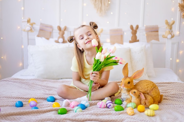 明るい色の卵とウサギが家で春の花のチューリップを持って明るい部屋で休日の準備をして笑顔で楽しんでいる小さな女の子をイースター