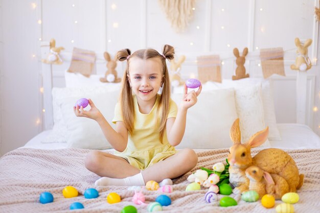 easter a little girl with painted colored eggs tulips and a rabbit at home in a bright room is preparing for the holiday smiling having fun and playing with eggs closing her eyes