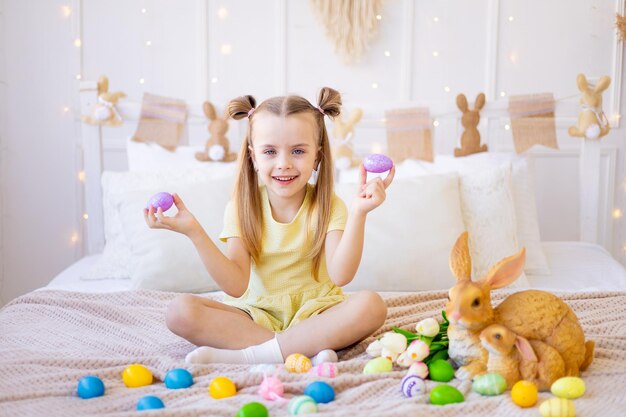 Easter a little girl with painted colored eggs tulips and a rabbit at home in a bright room is preparing for the holiday smiling having fun and playing with eggs closing her eyes