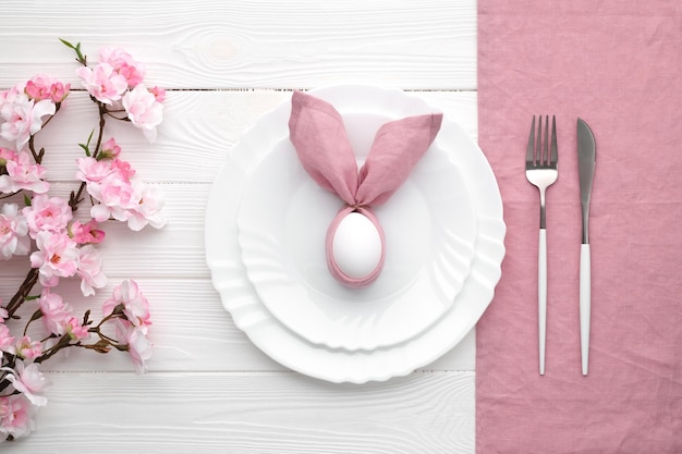 Photo easter lunch invitation festive table setting with pink spring flowers and cutlery bunny made of linen napkin and egg happy easter holiday menu concept empty plates white wooden background