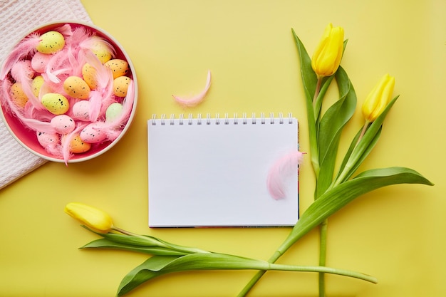 Easter mock up notepad Pink bright bowl with colorful eggs pink feathers and yellow tulips Happy Easter concept Post card mock up on yellow background