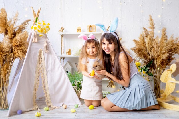 La mamma e la figlia di pasqua sorridono con un coniglio e le uova di pasqua e si preparano per la pasqua a casa in una stanza luminosa nelle orecchie di una lepre