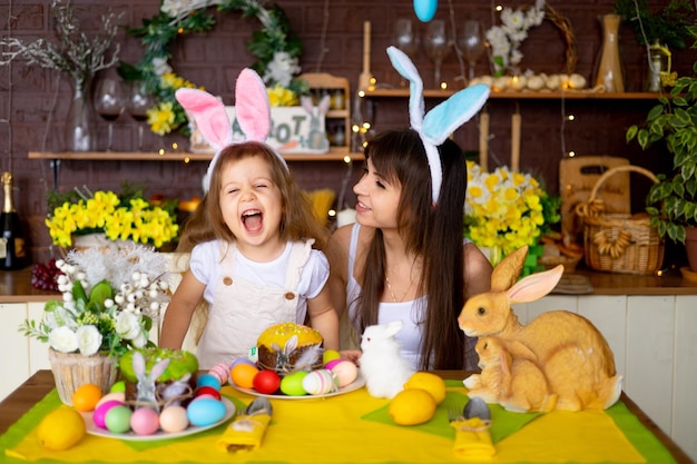 Easter mom and daughter with Easter eggs and cake at the festive table are smiling and preparing for Easter at home in the kitchen in the ears of a hare