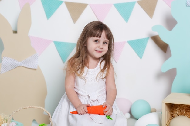 Easter! Portrait of a beautiful little girl in a white dress holding a carrot. Easter interior decoration, spring decor. Family holidays, traditions. Little farmer. Harvesting. Vegetarian, vegetables