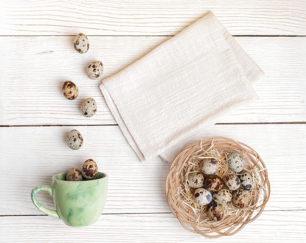 Easter quail eggs on woven plate eggs falling to green cup cotton napkin on white wooden backdrop