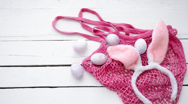 Easter still life with a pink string bag, decorative Easter bunny ears and eggs on wooden surface. Easter holiday concept.