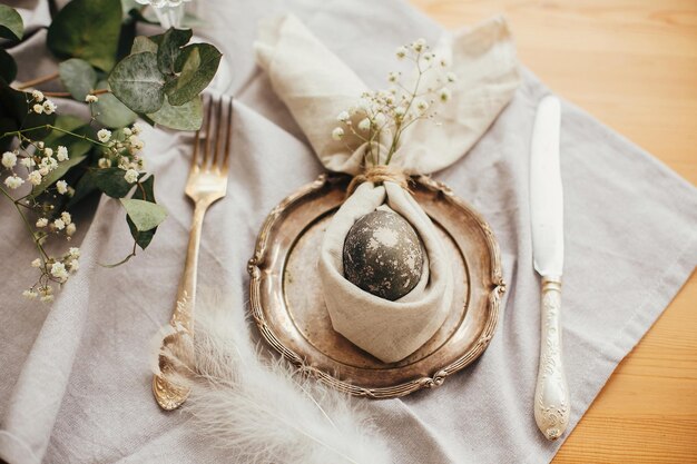 Easter table decorations modern natural dyed gray egg on napkin\
with bunny ears flowers on vintage plate and cutlery stylish easter\
brunch table setting with egg in easter bunny napkin