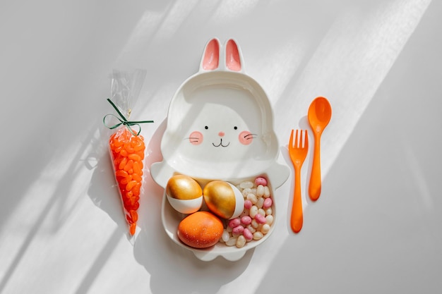 Украшение пасхального стола с милой тарелкой в форме кролика с пасхальными золотыми яйцами, конфетами и морковью Концепция счастливой Пасхи Идея для пасхального ужина