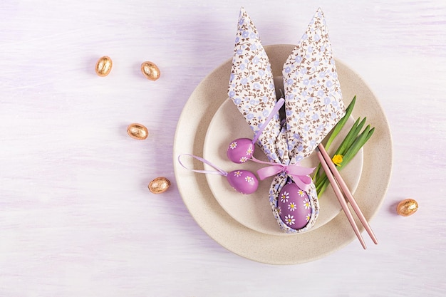 Easter table setting White plate with a napkin folded in the shape of a rabbit Easter and chocolate eggs on a pink background Happy Easter holiday concept for cafes and restaurants Top view flat lay