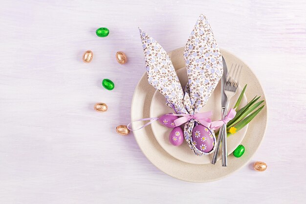 Easter table setting White plate with a napkin folded in the shape of a rabbit Easter and chocolate eggs on a pink background Happy Easter holiday concept Top view flat lay