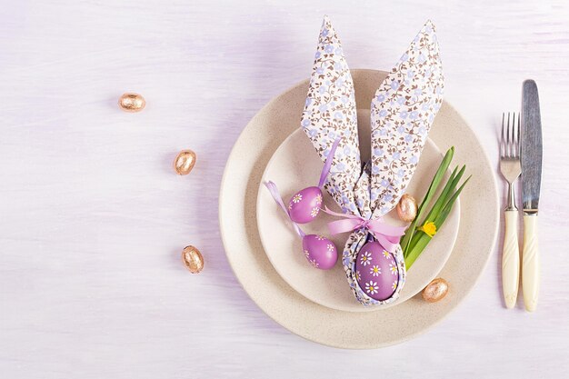 Easter table setting White plate with a napkin folded in the shape of a rabbit Easter and chocolate eggs on a pink background Happy Easter holiday concept Top view flat lay