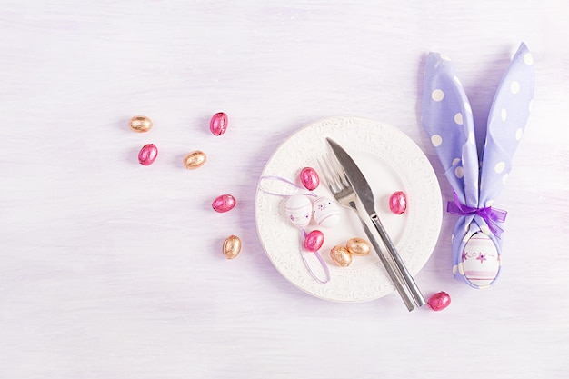 Easter table setting White plate with a napkin folded in the shape of a rabbit Easter and chocolate eggs on a pink background Happy Easter holiday concept Top view flat lay