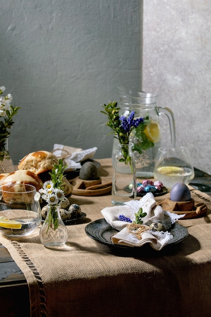 Photo easter table setting with colored and chocolate eggs, hot cross buns, bouquet flowers, empty ceramic plate with napkin, glass of lemonade drink on wooden table with textile tablecloth