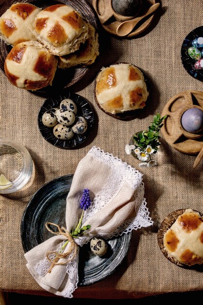 Easter table setting with colored and chocolate eggs, hot cross buns, bouquet flowers, empty ceramic plate with napkin,