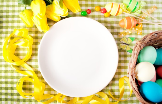 Foto regolazione della tavola di pasqua con piatto bianco, cesto di uova colorate, candele e tulipani gialli.