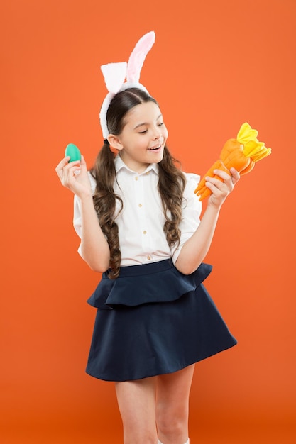 うさぎの耳のイースターの雰囲気の子供はニンジンを食べるのが大好き 子供のバニーの衣装 ニンジンで小さな幸せな女の子 塗装された卵を保持します エッグハントは健康な子供時代を始めました うさぎのようにニンジンをかじります