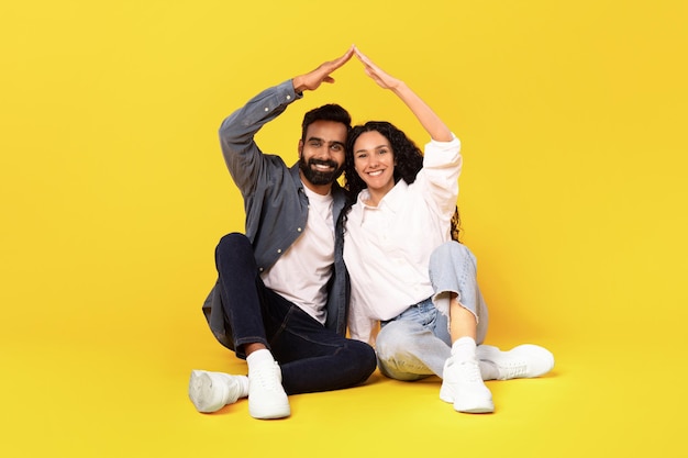 Eastern couple making roof joining hands sitting over yellow background