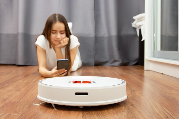 Easy cleaningYoung European girl resting with smartphone while robot vacuum cleaner collects dust