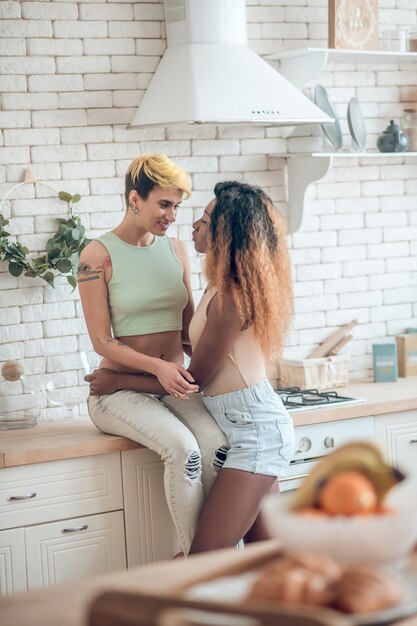 簡単なコミュニケーション。台所のテーブルに座っている笑顔の短い髪のガールフレンドの近くに立っているショートパンツの長い髪の若いアメリカ人女性