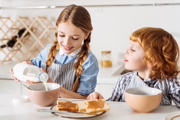 Easy cooking. Beautiful admirable lovely child sharing a morning meal with her brother and poring milk in her bowl while they both sitting at the table in a kitchen