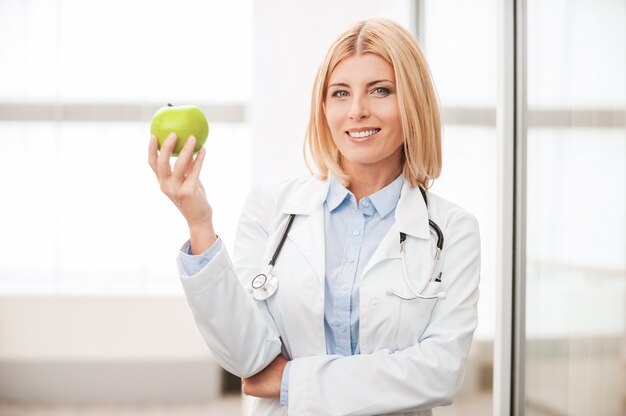 Mangiare sano! fiduciosa dottoressa in uniforme bianca che tiene in mano una mela verde e sorride mentre si appoggia alla parete di vetro