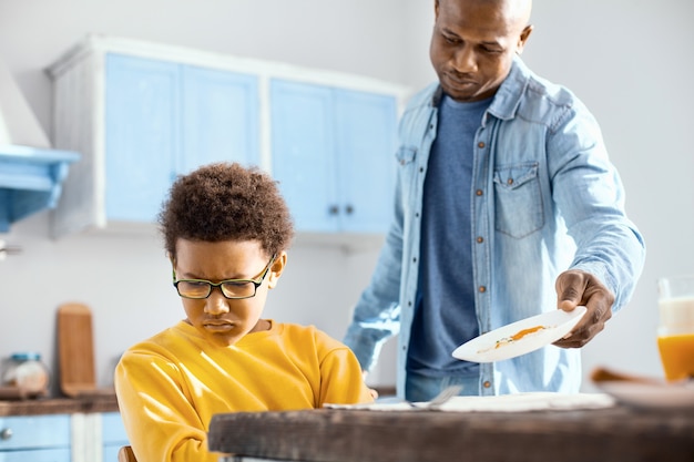 Mangia per favore. amorevole giovane padre che chiede a suo figlio di fare colazione mentre il ragazzo gira la faccia dall'altra parte, mostrando la sua riluttanza