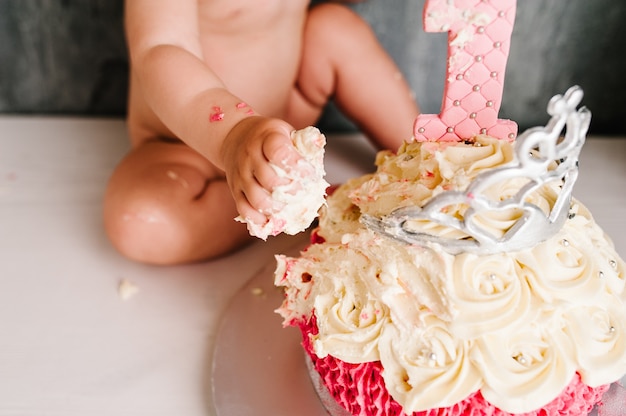 Mangiando le mani di torta. ritratto di una bambina caucasica che festeggia il suo primo compleanno con una torta bianca e rosa gourmet con candela 1 anno e corona sopra. torta smash primo anno concetto.