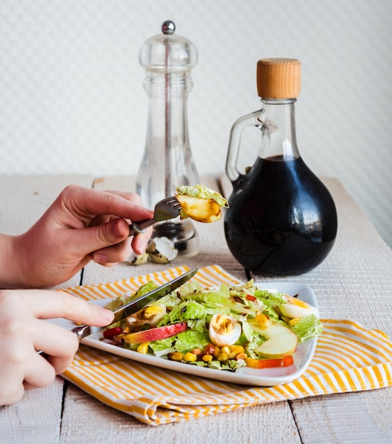 Есть зеленый салат с перепелиными яйцами, кукурузой и яблоком, рука, вилка и нож