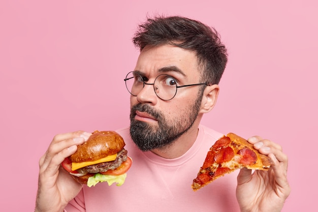 Eating junk food. Serious attentive bearded adult European man holds tasty hamburger and slice of pizza wears spectacles eats cheat meal 