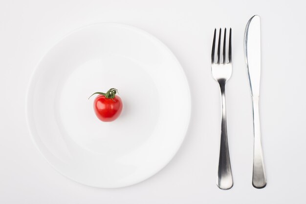 Eating on a low calorie concept. Top above overhead view photo of a plate with a single tomato with fork and knife placed to the right side isolated on white background