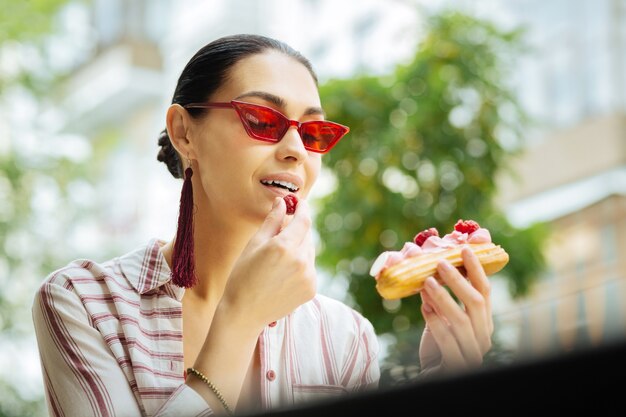 Eating raspberries. Cheerful beautiful visitor of a cafe holding a delicious eclair and eating raspberries on the top of it