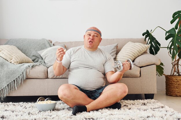 Photo eats and does exercises funny overweight man in casual clothes is indoors at home