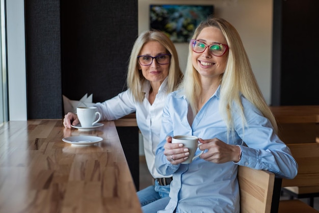 Eautiful blonde with glasses and her elderly mother are sitting in a cafe and drinking coffee Mothers Day Beautiful aged woman and her adult daughter are smiling and chatting Related resemblance