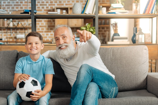 Echte mannen. Gelukkig jongetje met een bal en kijken naar een voetbalwedstrijd op tv samen met zijn grootvader wijzend op het scherm