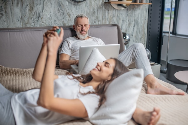 Echtpaar. Ernstige attente grijsharige man aan het werk op laptop en vrolijke jonge vrouw met smartphone liggend in de slaapkamer