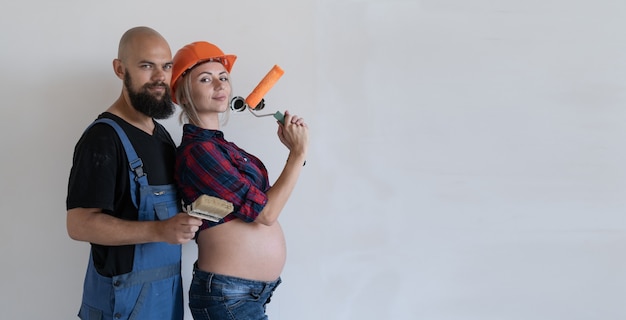 Echtpaar man en vrouw gekleed in werkkleding staan in de buurt van een witte muur. zwanger meisje. we doen onze eigen kopieerruimte voor huisrenovatie.